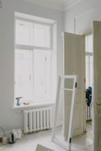 A person examines paint layers on a wooden window frame in a sunlit, older home’s interior.
