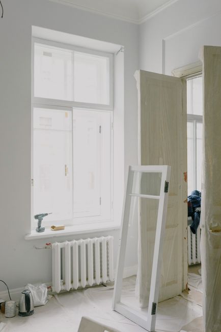 A person examines paint layers on a wooden window frame in a sunlit, older home’s interior.